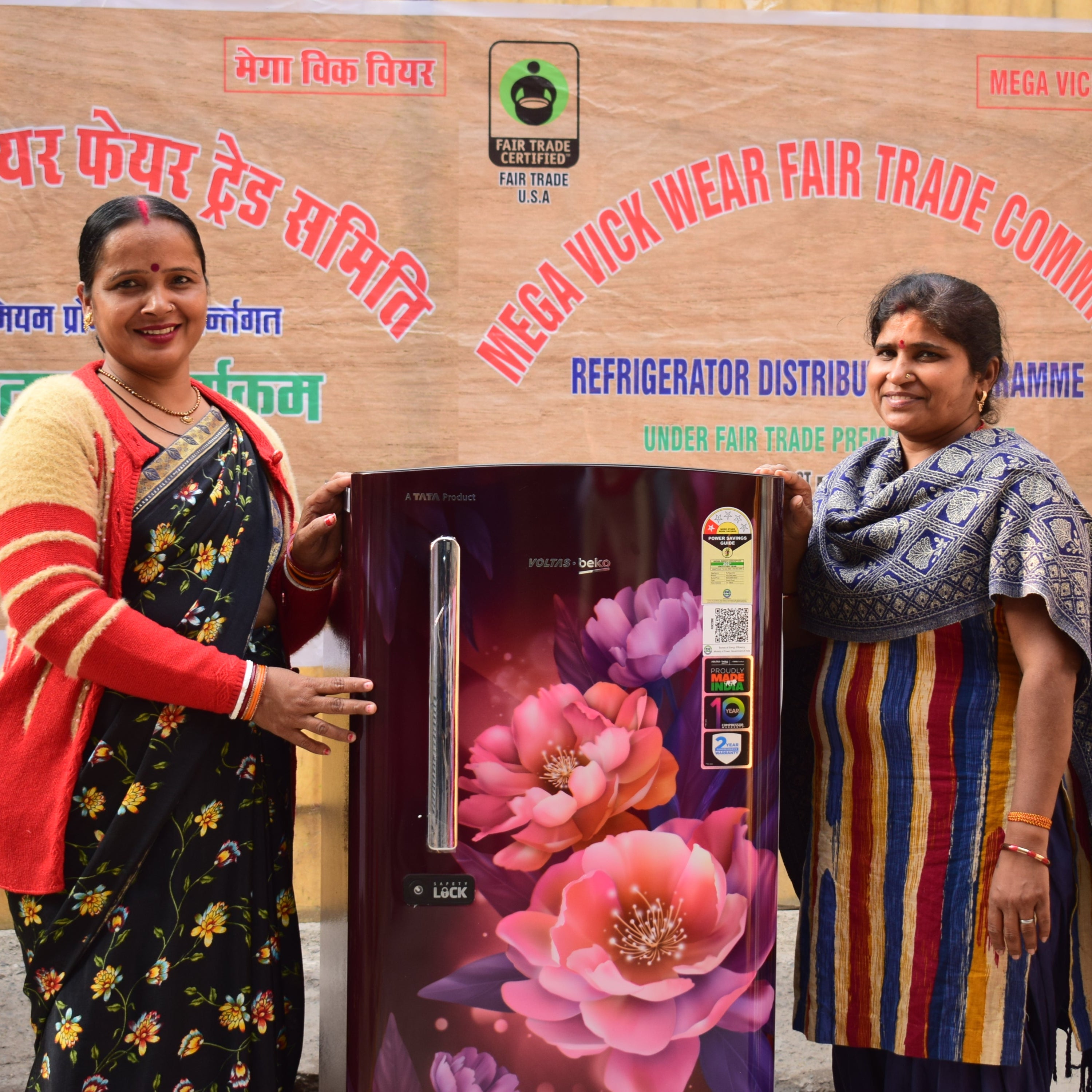 Two women holding a refrigerator with a large 'Mega Vick Wear Fair Trade Committee' sign in the background.